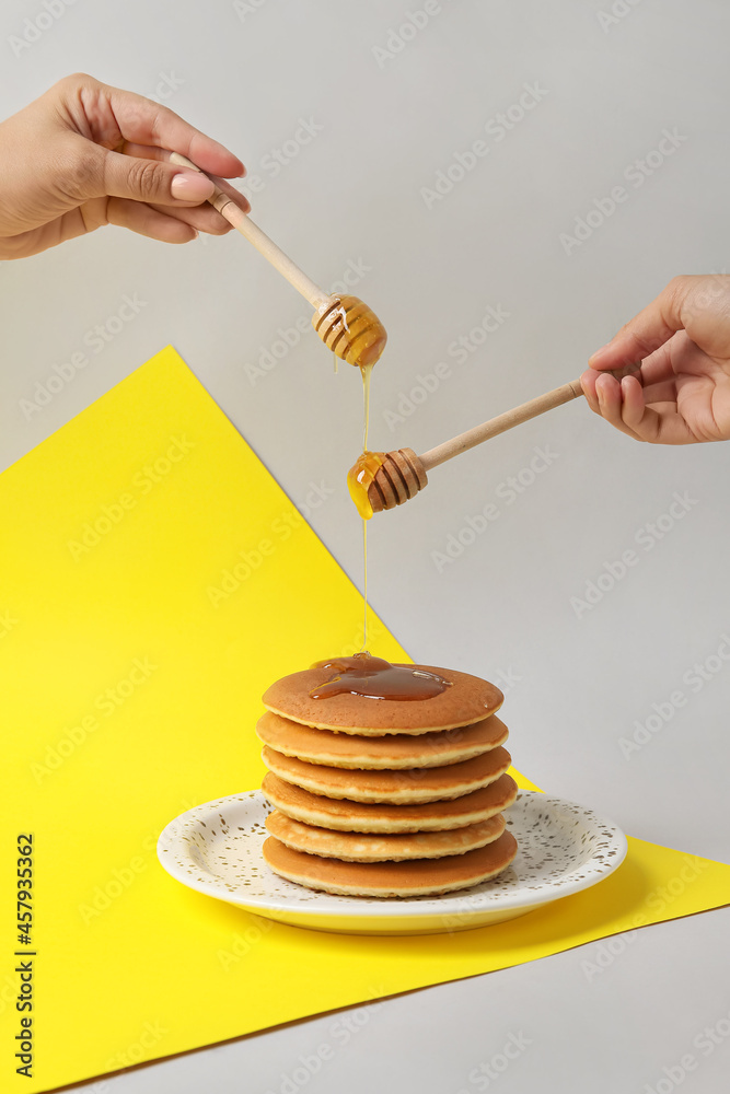 Female hands pouring honey onto plate with tasty pancakes on color background