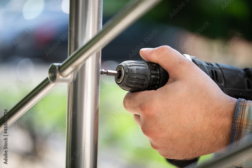 A worker in gloves repairs a metal railing of a staircase using a screwdriver and a drill