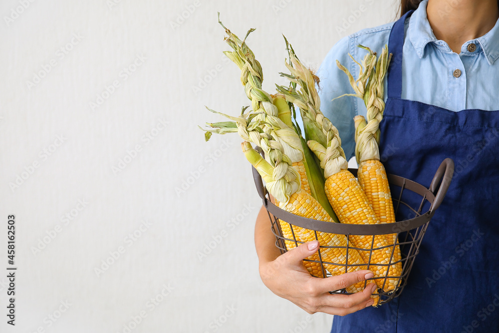 Woman holding basket with fresh corn cobs on light background