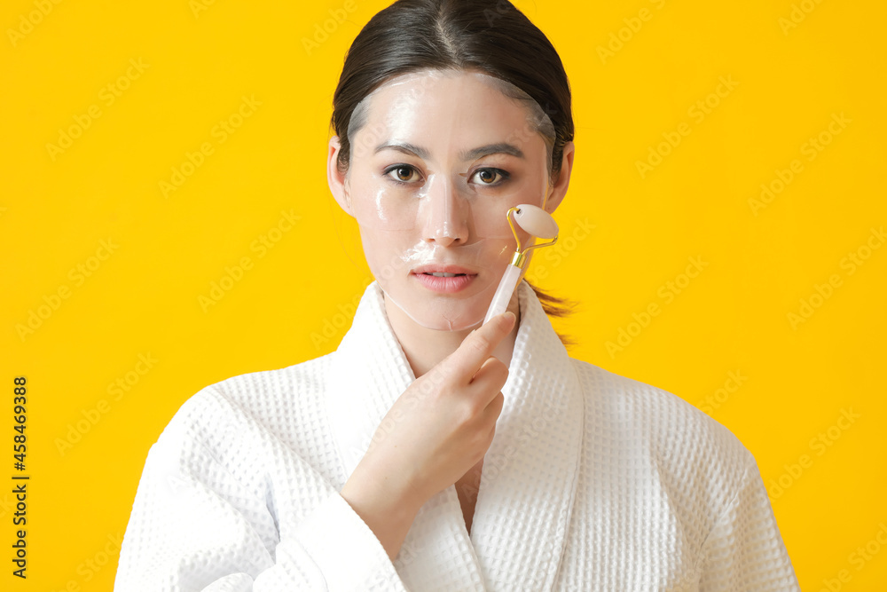 Beautiful young Asian woman with sheet facial mask and massage tool on color background