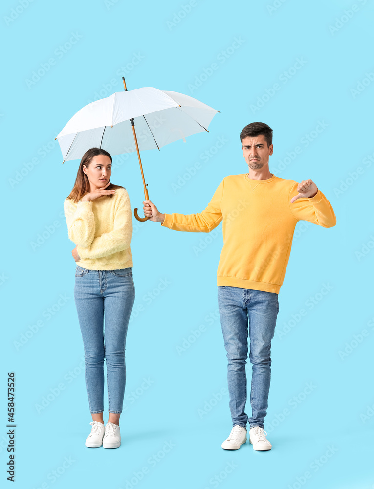 Displeased guy sharing umbrella with young woman on color background