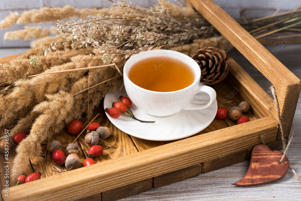 Ceramic cup of tea with saucer, rosehip berries, acorns and dried plants on a wooden tray. Autumn mo