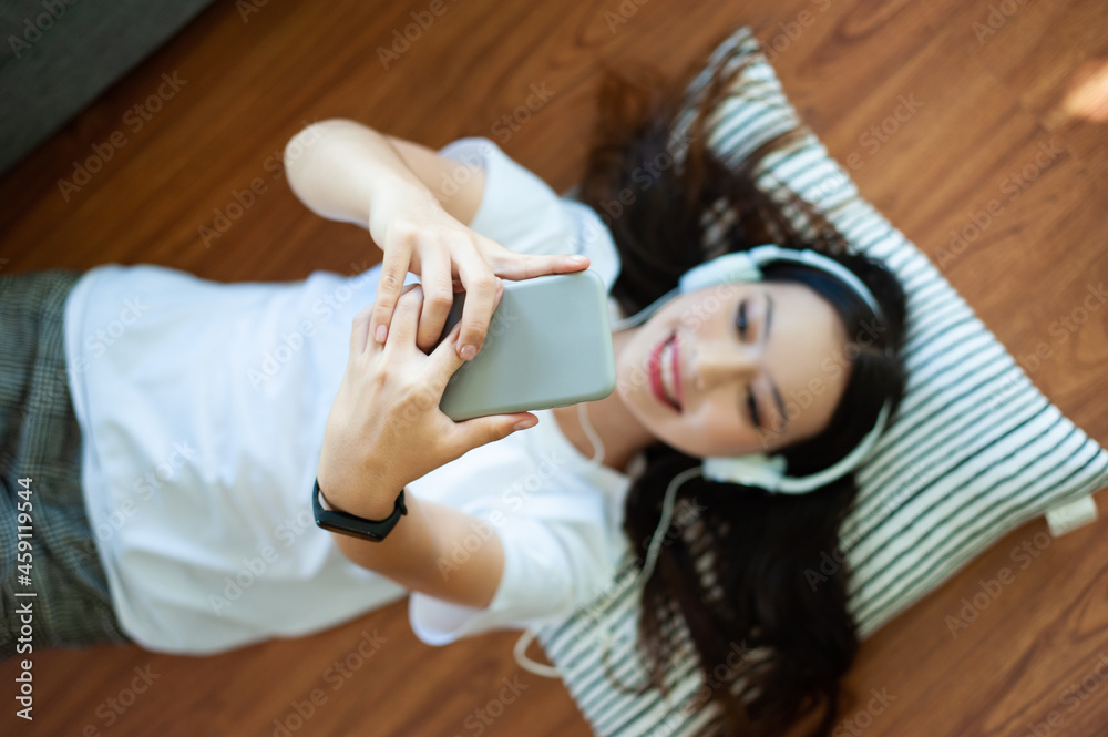 Young woman listening to music with headphones playing mobile phone while lying on the floor in livi