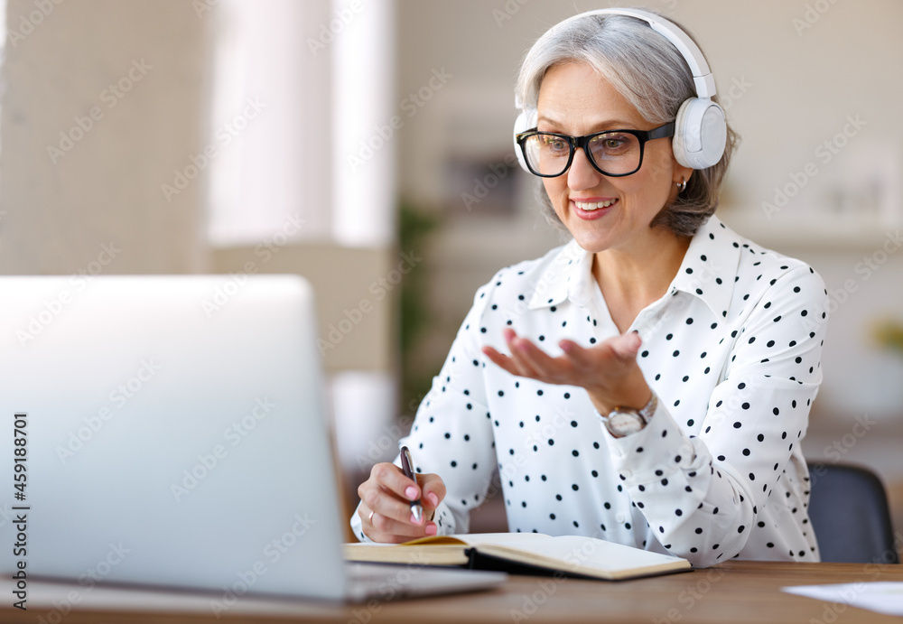 Beautiful smiling mature woman in headphones during online meeting on laptop with colleagues