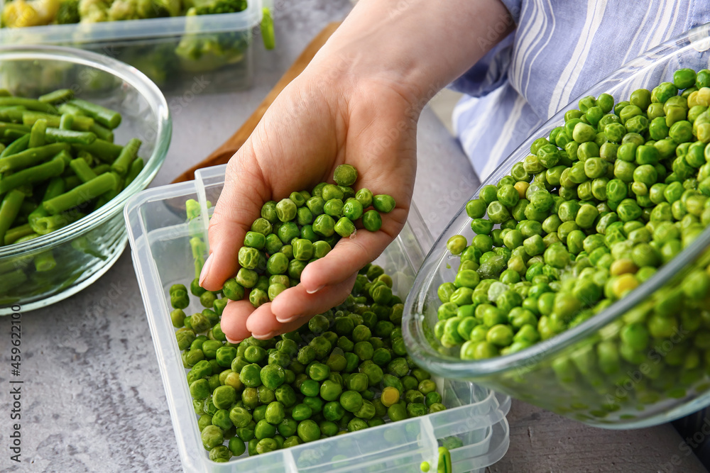 Woman putting frozen peas in plastic container at table in kitchen, closeup