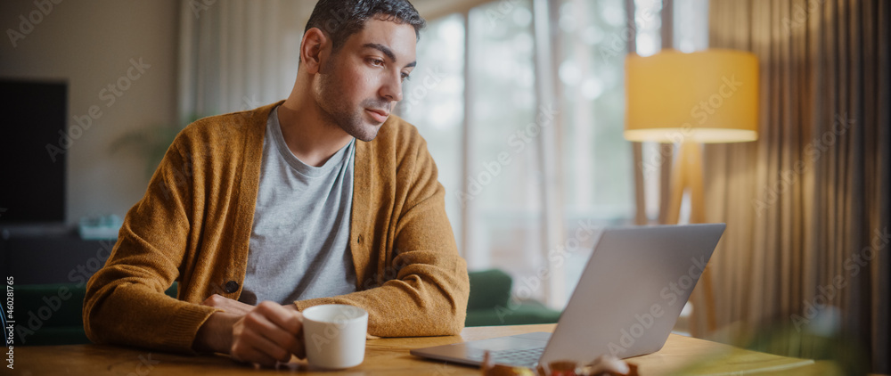 Handsome Adult Man Using Laptop Computer, Sitting in Living Room and Drinking Tea or Coffee in Apart