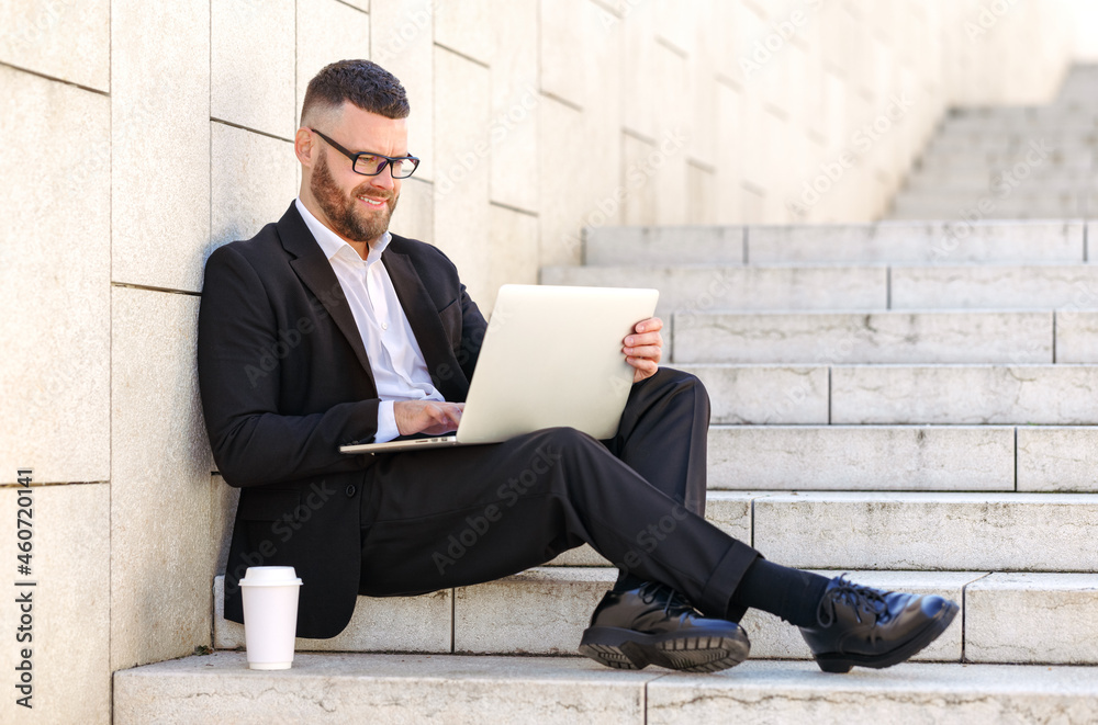 Young happy businessman freelancer in suit sitting on stairs outside with laptop and coffee to go