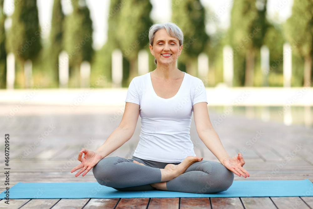 Calm happy senior woman sitting in lotus pose on mat during morning meditation in park