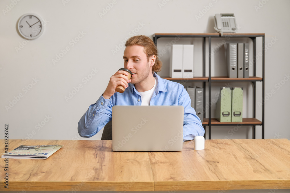 Handsome young man with laptop drinking coffee in office