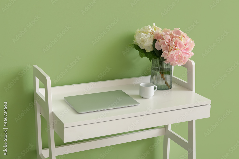 Vase with hydrangea flowers and laptop on table near color wall