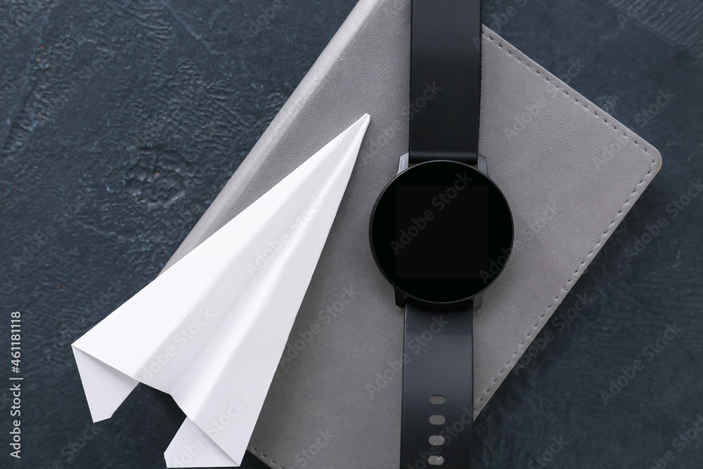 Paper airplane, wrist watch and notebook on dark background, closeup