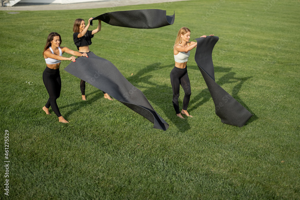 European girlfriends putting fitness mats on green meadow ​before sports or yoga training at sunny d