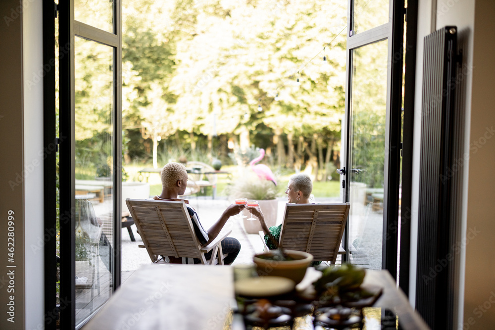 Multiracial couple drinking cocktails while sitting on wooden chairs at home terrace. European girl 