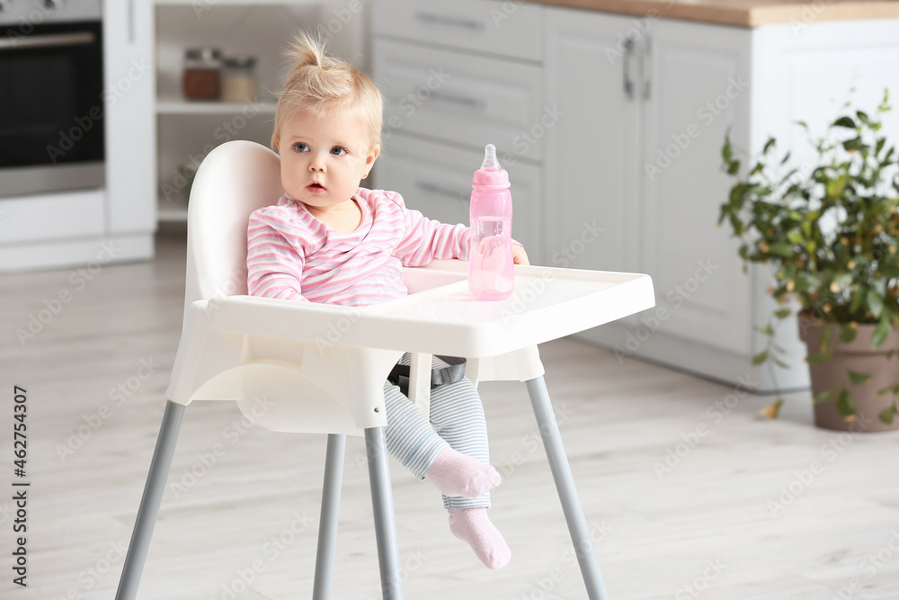 Cute baby girl with bottle of water sitting on high chair in kitchen