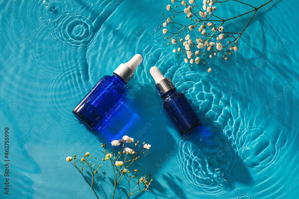 Cosmetic dropper bottles and flowers in water on color background