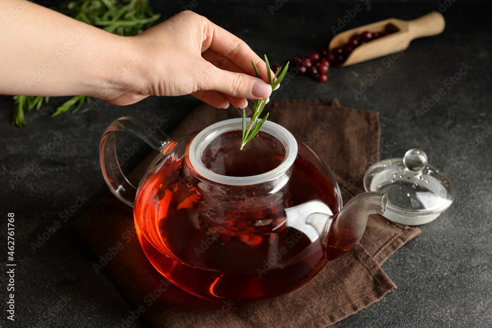 Woman putting rosemary into teapot with cranberry tea on dark background