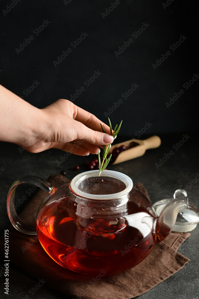 Woman putting rosemary into teapot with cranberry tea on dark background, closeup