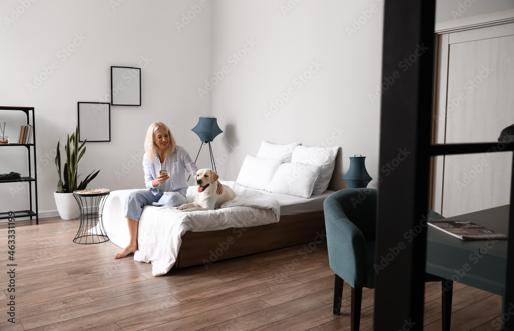 Mature woman with cute Labrador dog using mobile phone in bedroom