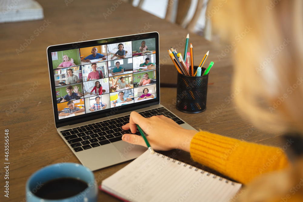 Caucasian woman using laptop for video call, with smiling diverse elementary school pupils on screen