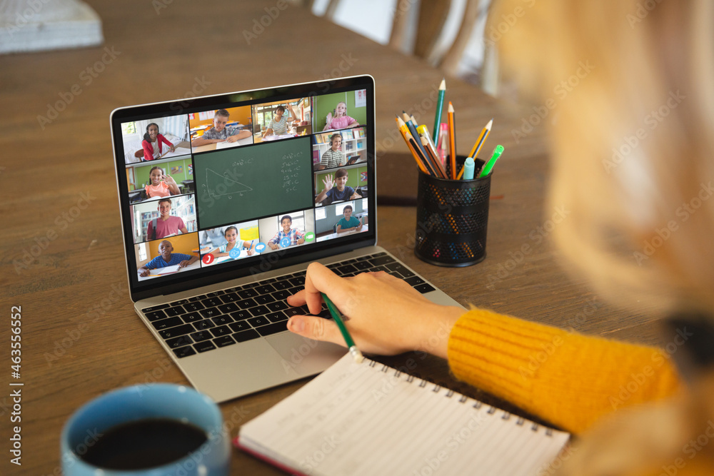 Caucasian woman using laptop for video call, with smiling diverse elementary school pupils on screen