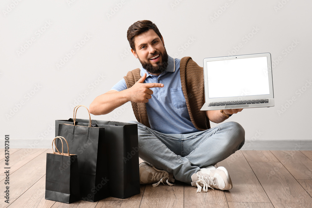 Handsome man with Black Friday shopping bags pointing at laptop near light wall