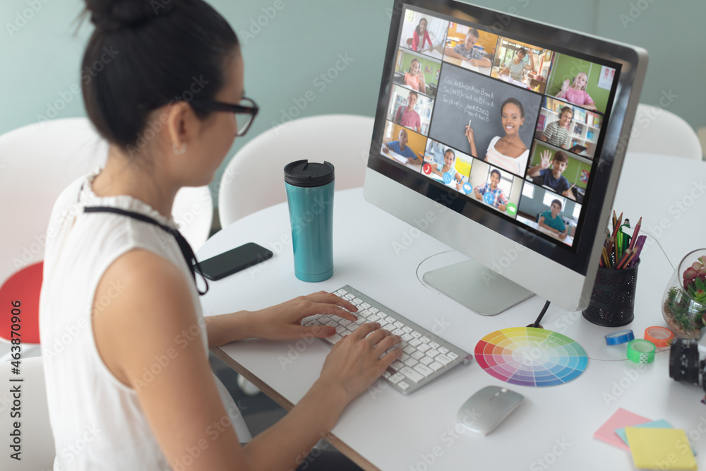 Asian woman using computer for video call, with diverse elementary school pupils on screen