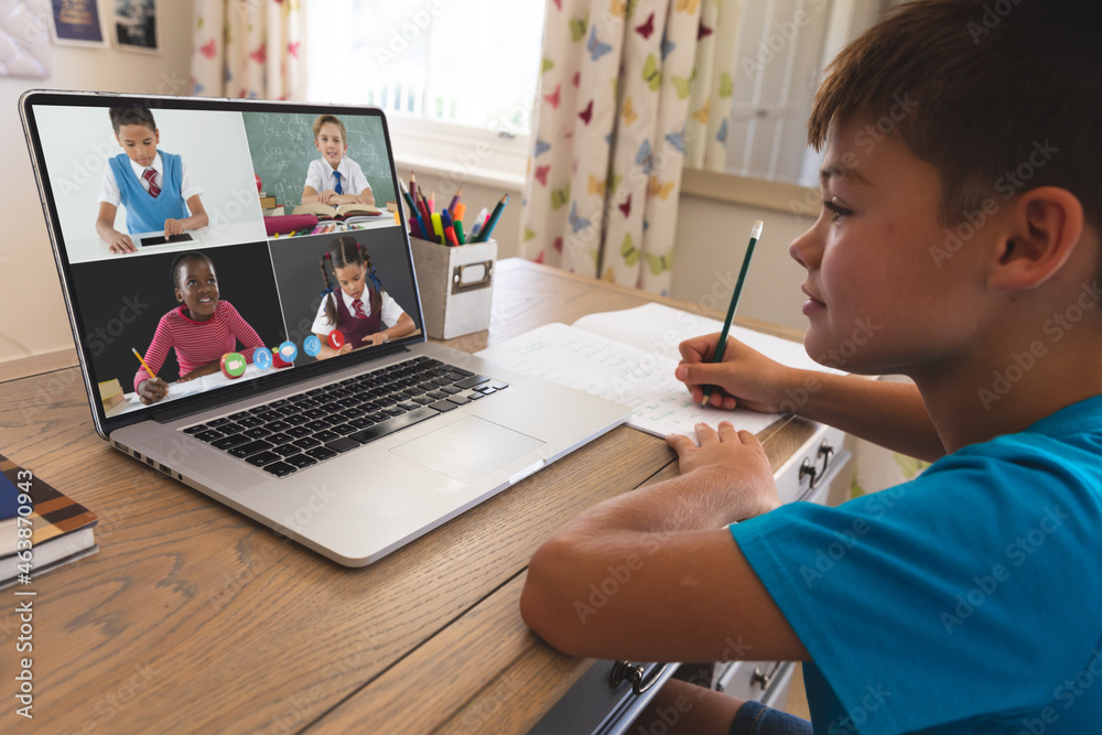 Smiling caucasian boy using laptop for video call, with diverse elementary school pupils on screen