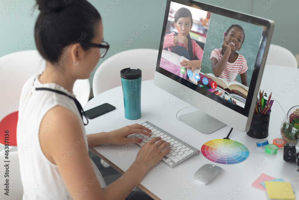 Asian woman using computer for video call, with smiling diverse elementary school pupils on screen