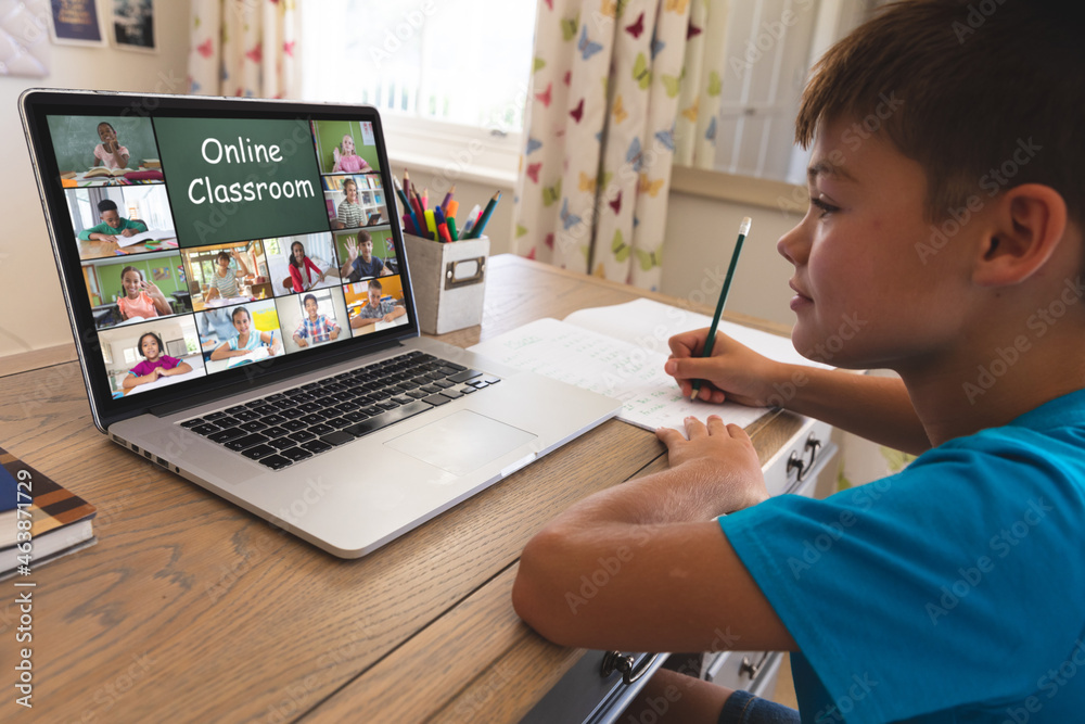 Caucasian boy using laptop for video call, with smiling diverse elementary school pupils on screen