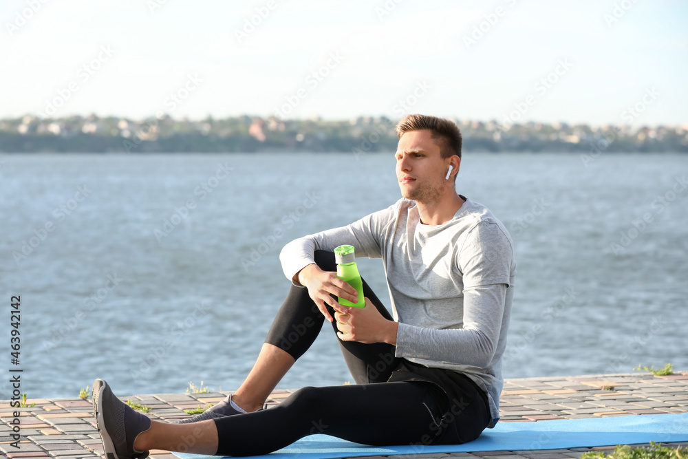 Sporty young man with bottle of water sitting near river