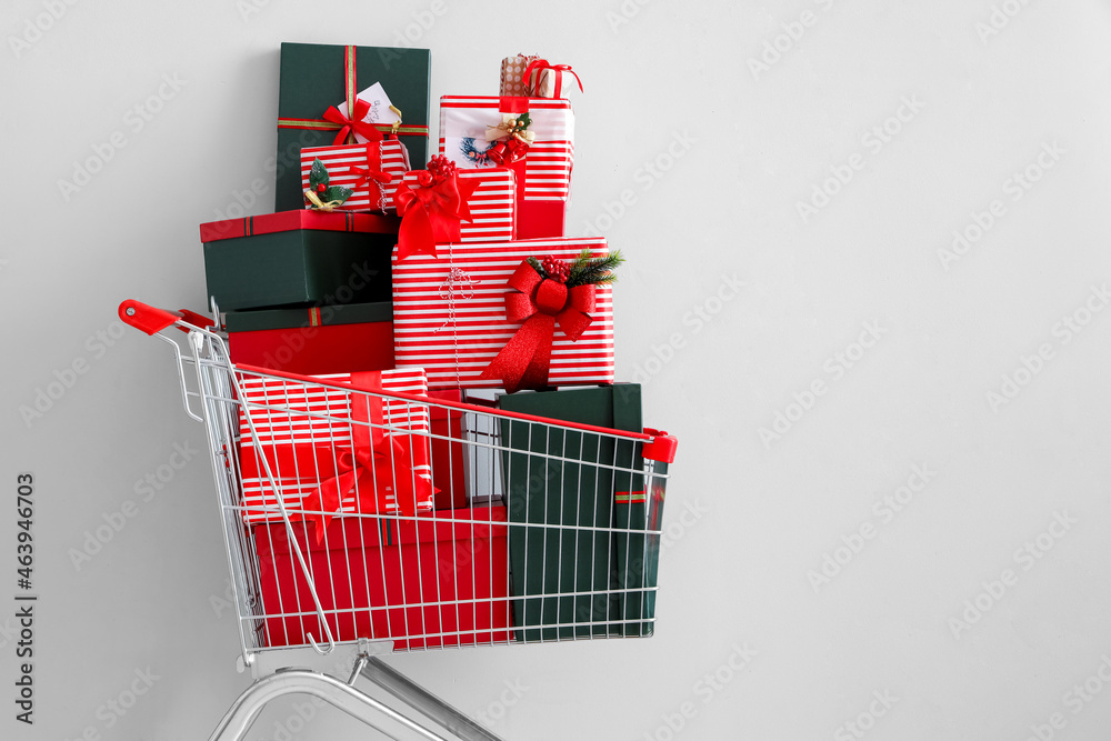 Shopping cart with beautiful Christmas gifts near light wall