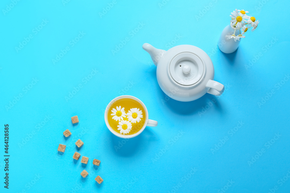 Teapot and cup of natural chamomile tea with flowers on blue background