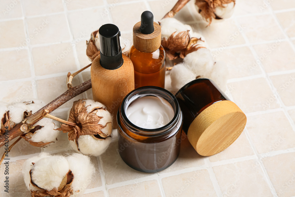 Different cosmetic products and beautiful cotton branch on light tile, closeup