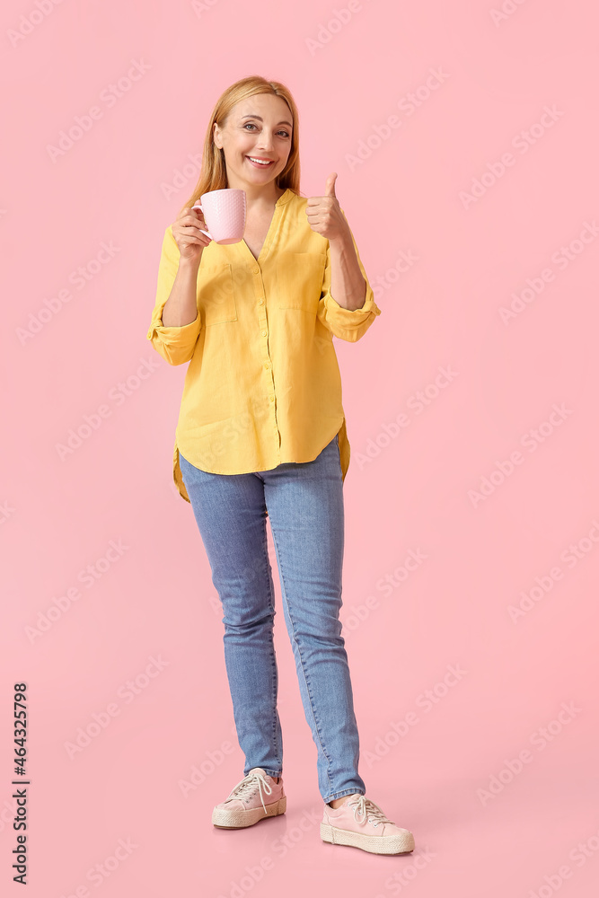 Beautiful mature woman with cup of coffee showing thumb-up on color background