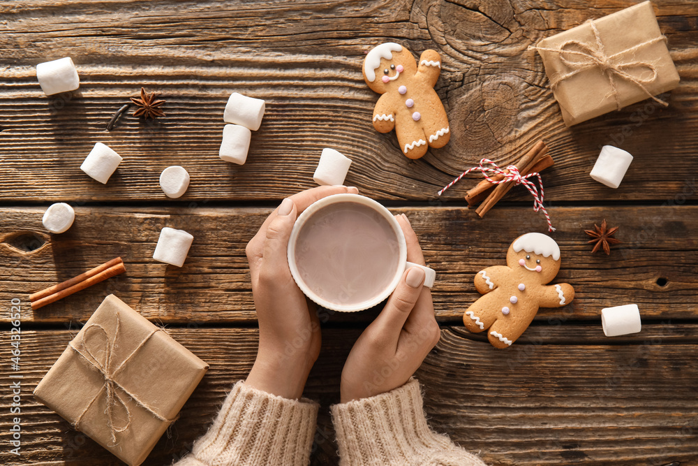 Female hands with cup of cacao, marshmallows, cookies and gifts on wooden background