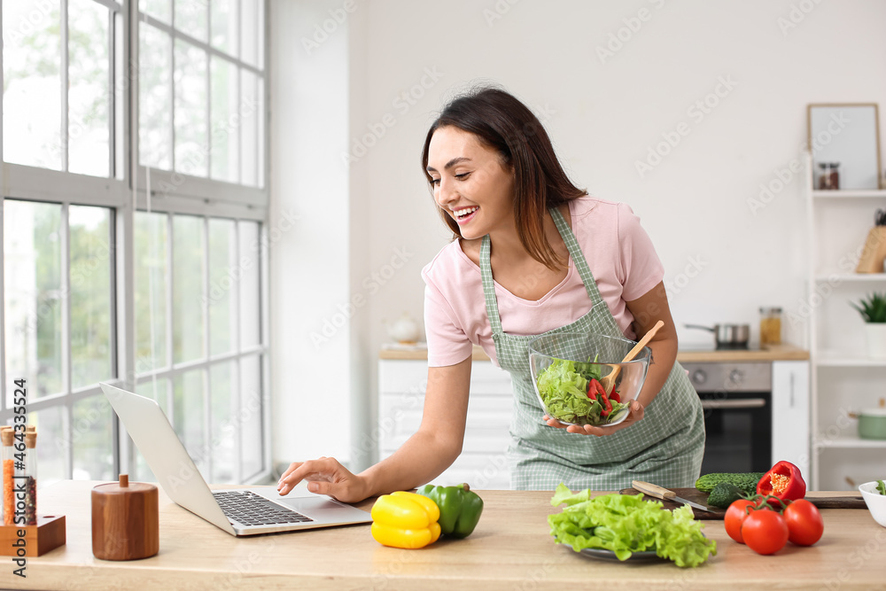 Young woman with fresh vegetable salad and laptop in kitchen