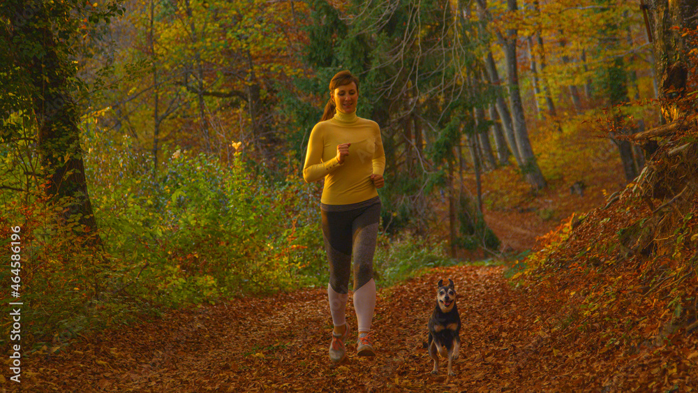 Woman looks down at her dog while jogging down the scenic forest colored trail.