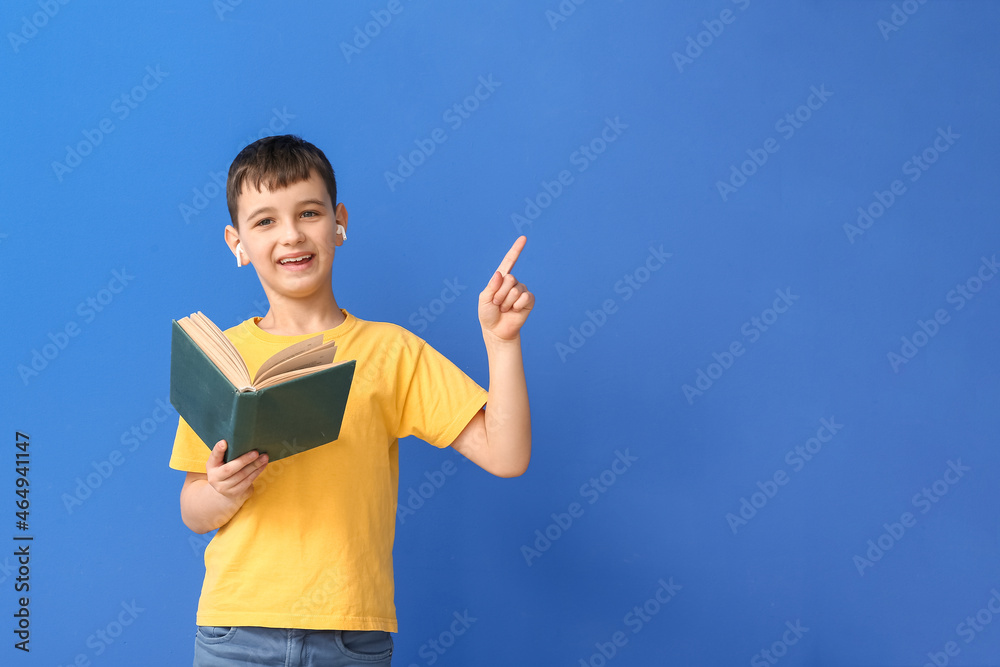 Little boy with book and earphones pointing at something on color background