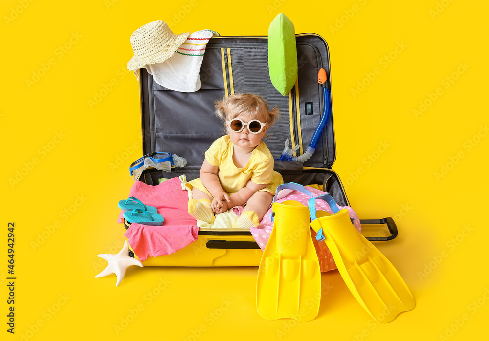 Cute baby girl in suitcase with belongings on color background