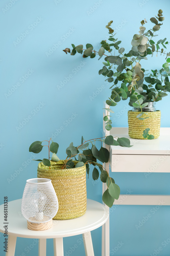 Baskets with eucalyptus branches and lamp on tables near blue wall