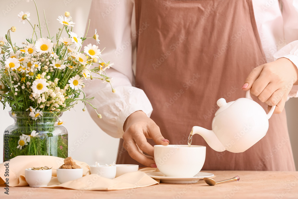 一名妇女将新鲜的洋甘菊茶从茶壶倒进桌子上的杯子里