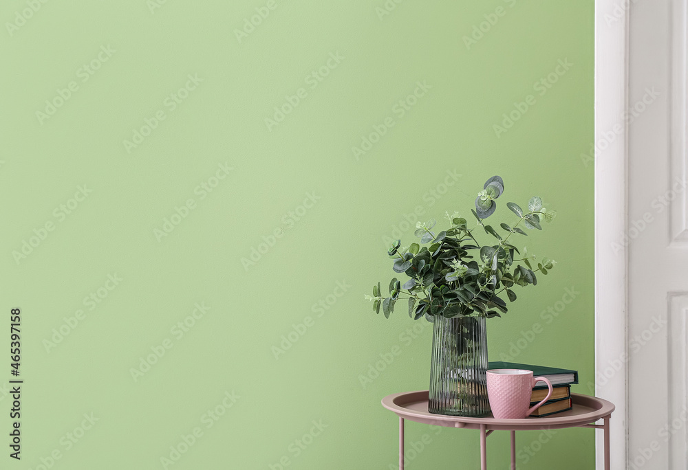 Vase with eucalyptus branches, cup of tea and books on table near color wall