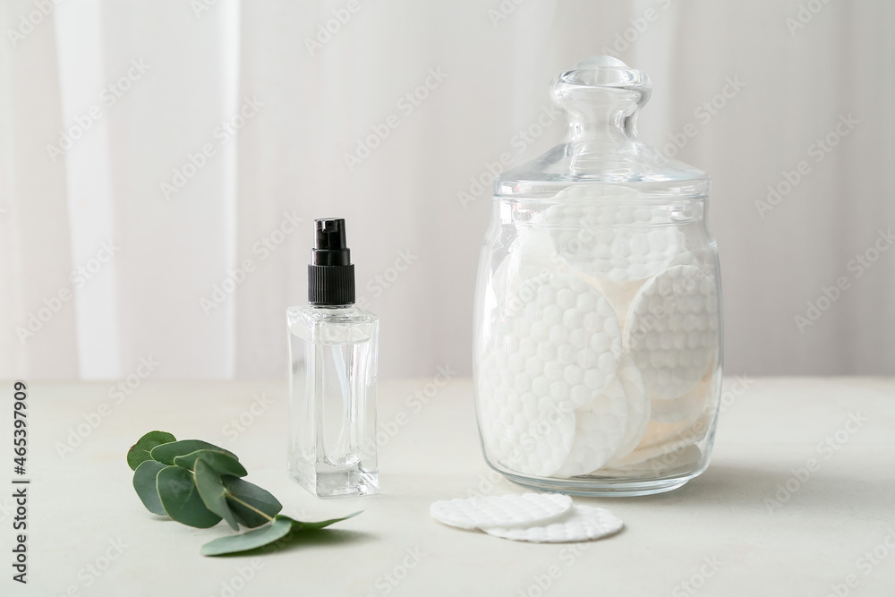 Glass jar with clean cotton pads and bottle of cosmetic product on light table