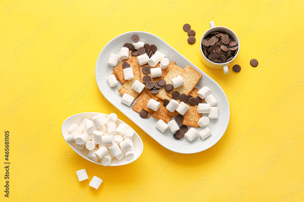 Plate with crackers, marshmallows and chocolate on yellow background