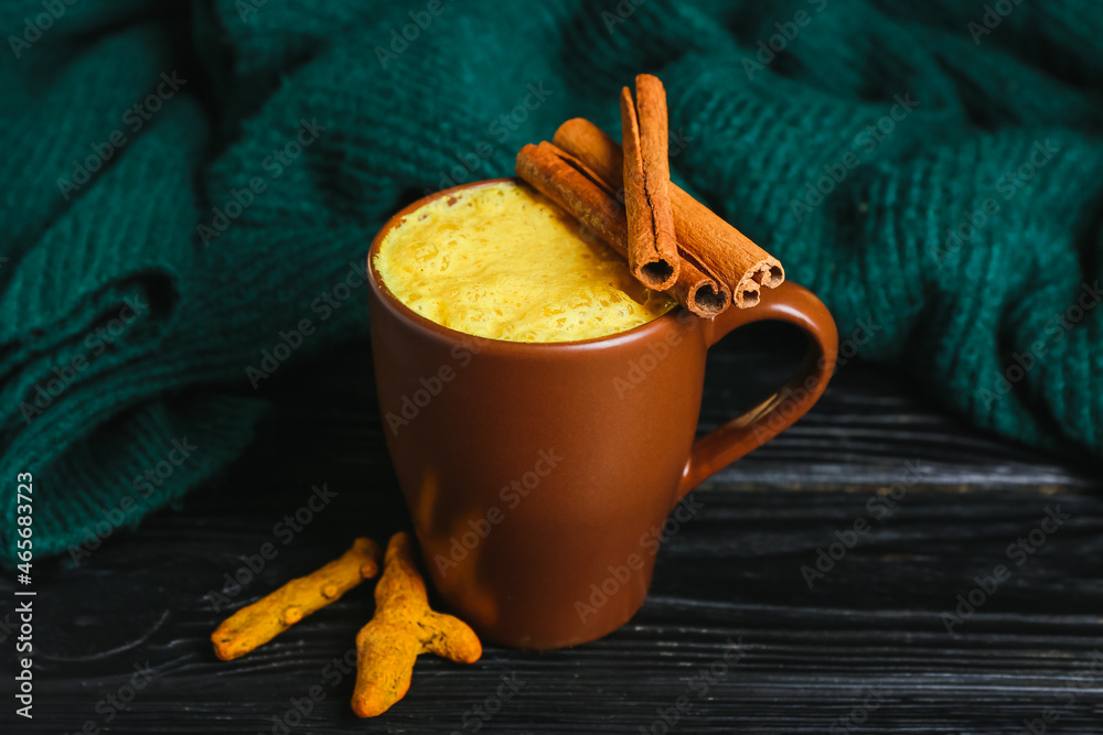 Cup of tasty turmeric latte with cinnamon and sweater on dark wooden background