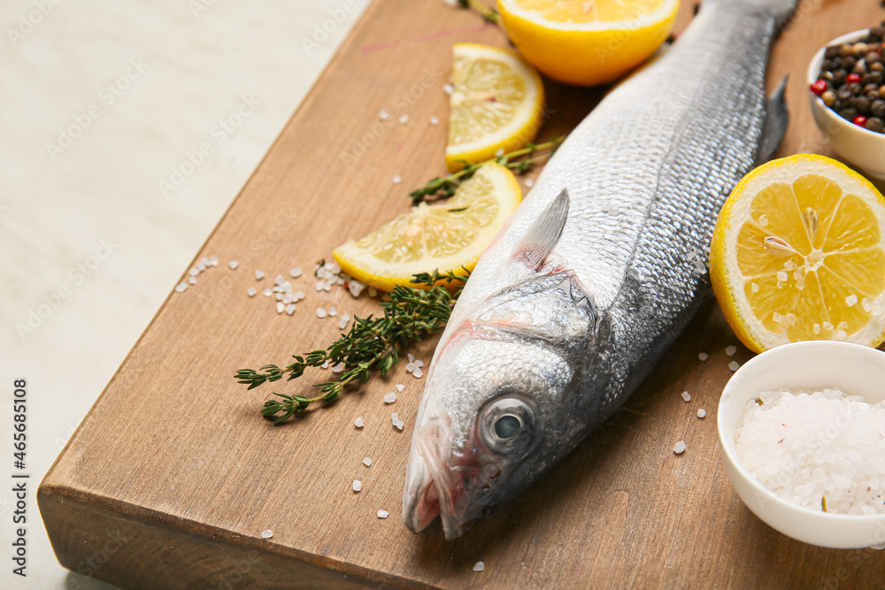 Wooden board with fresh uncooked sea bass fish, lemon and spices on white marble background