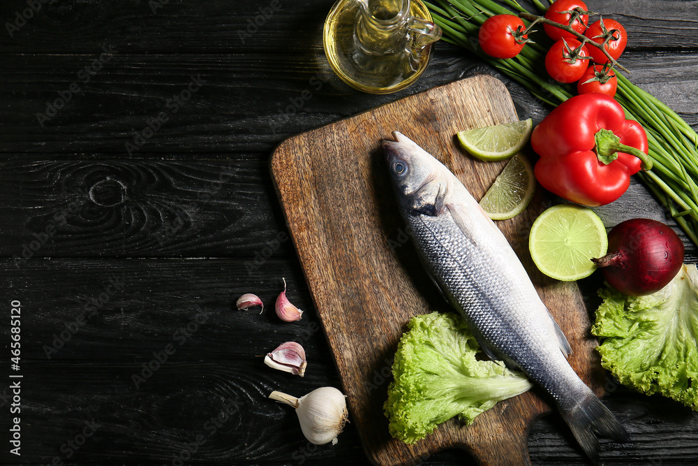 Board with fresh uncooked sea bass fish and vegetables on black wooden background