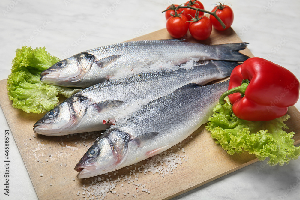 Board with fresh uncooked sea bass fish and vegetables on white marble background