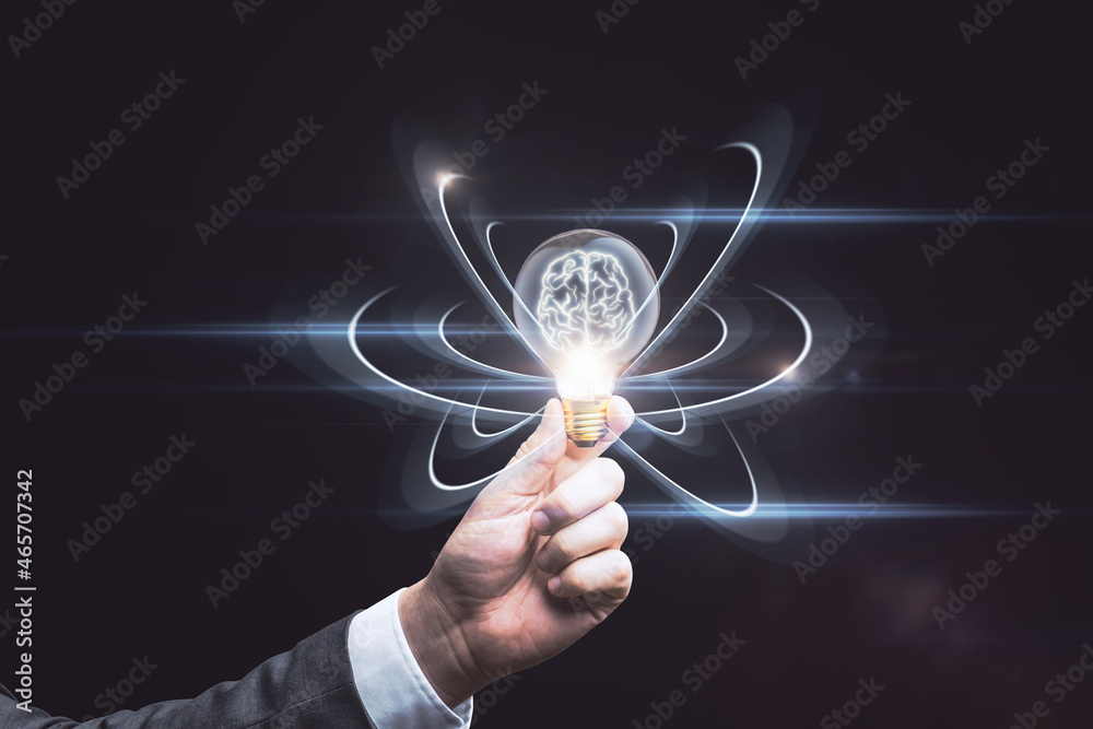 Close up of hand holding abstract light bulb on dark wide background with digital brain and atom out