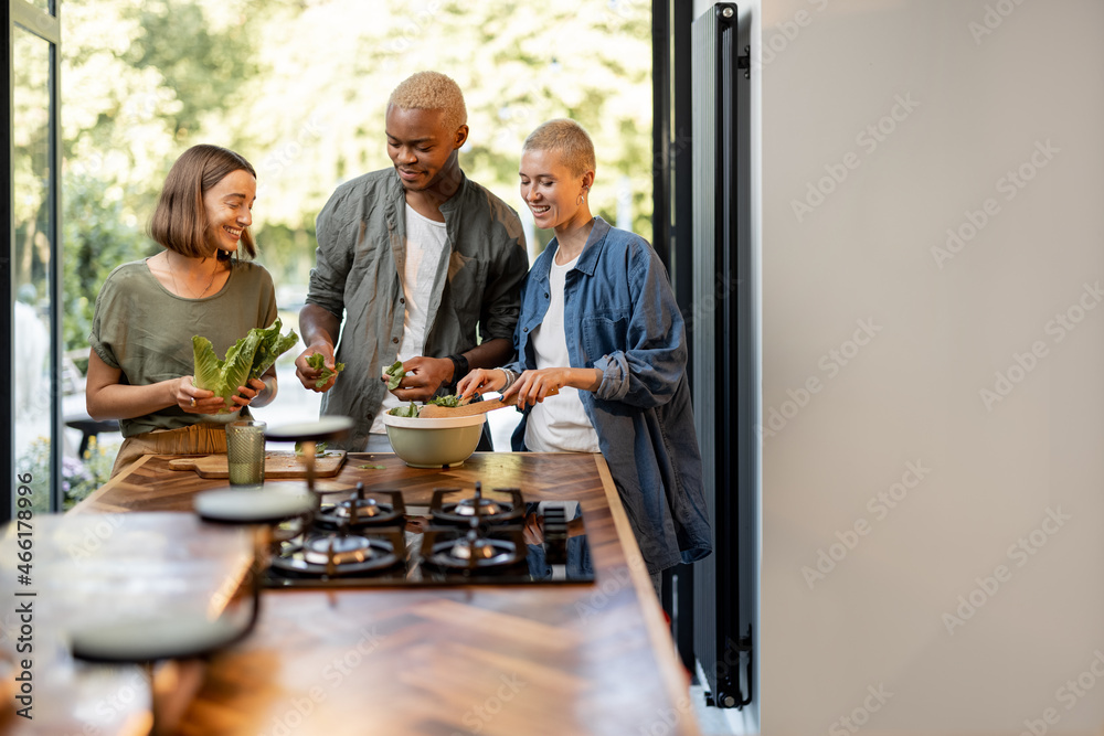 Friends cooking salad at home kitchen. Black man and european girls enjoying time together. Concept 